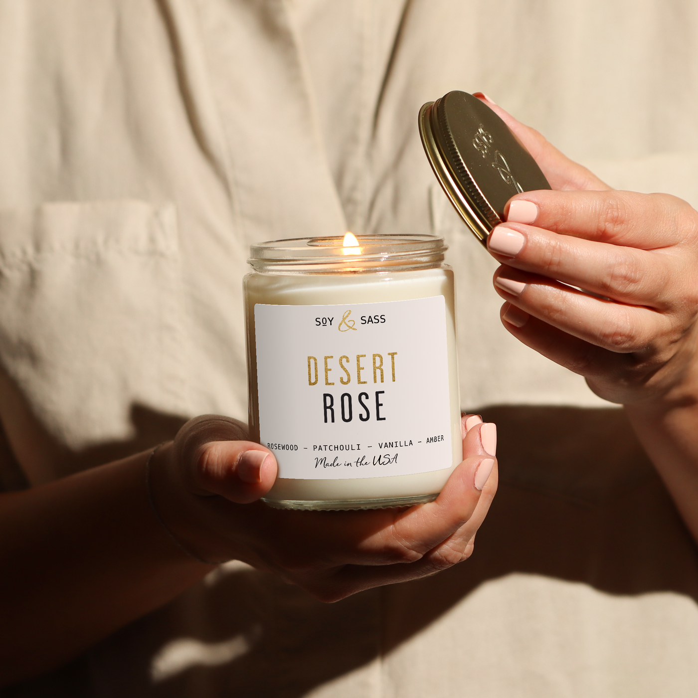 Person holding a 'Desert Rose' candle with a lit wick, wearing a light-colored shirt.
