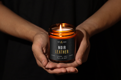 Hands holding a 'Noir Leather' candle against a dark background